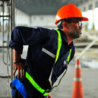 Un anciano con un casco naranja se sujeta la espalda con dolor mientras trabaja en una zona de construcción