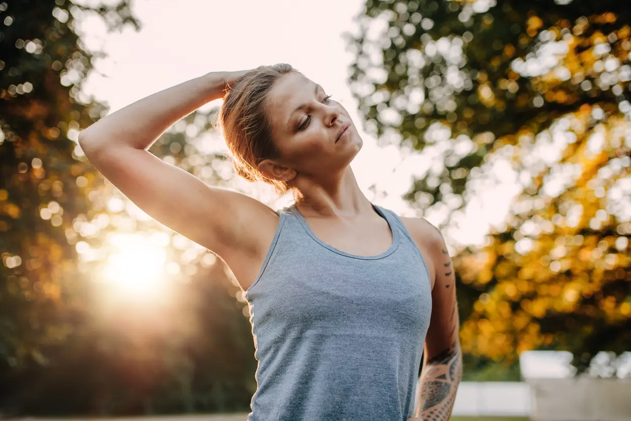 Portrait of healthy young woman stretching her neck outdoors. Caucasian fitness model warming up in park.