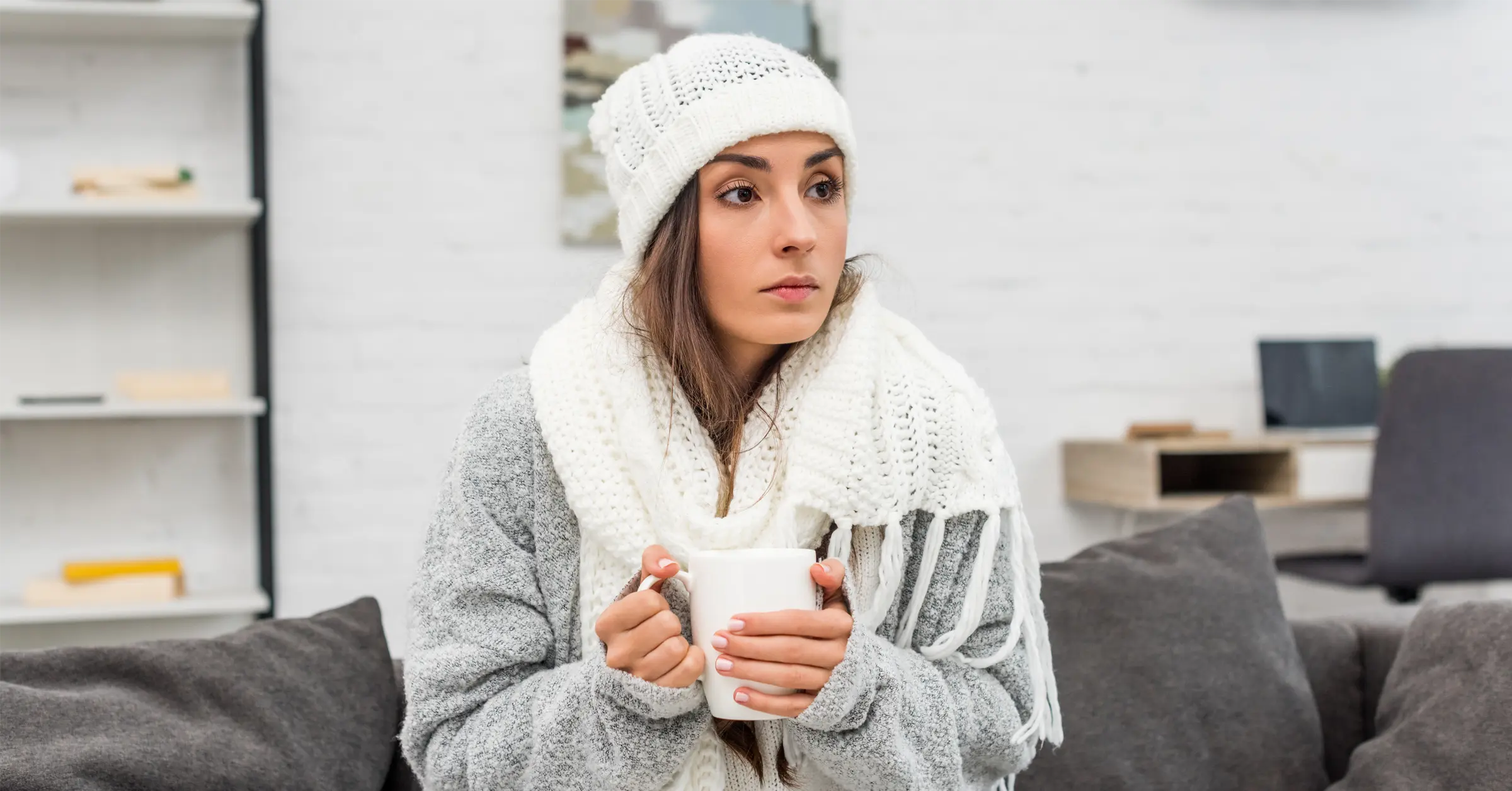 Young woman in warm clothes holding cup of tea while sitting on couch at home