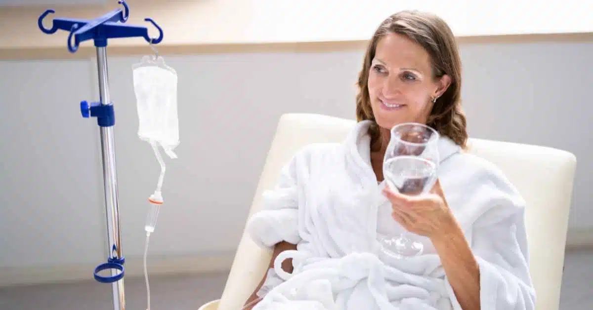 lady in medical clinic with IV drip drinking a glass of water, smiling