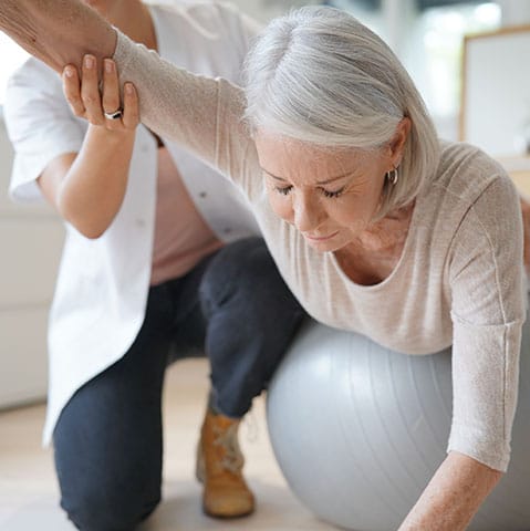 Older woman stretching arm out on a medicine ball with the help of a female physical therapist