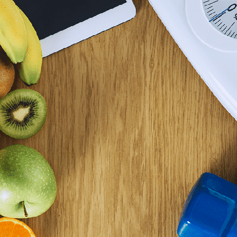 Fruit, ipad, scale, and a blue dumbell sitting on a wooden table for weight loss