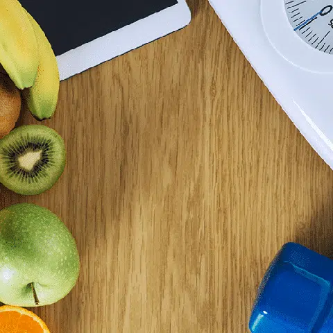 Fruit, ipad, scale, and a blue dumbell sitting on a wooden table for weight loss