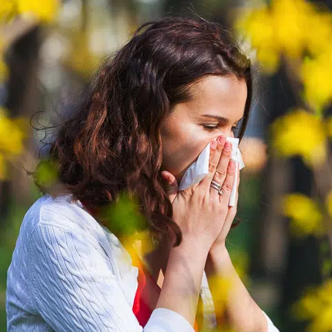Mujer estornudando por alergia con flores amarillas detrás de ella